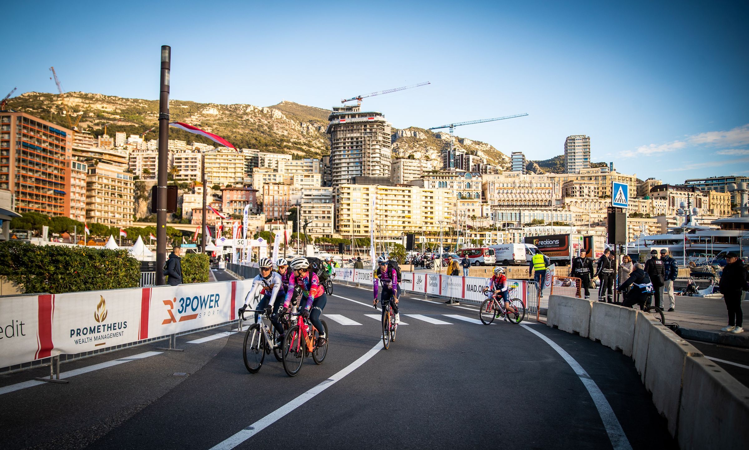 Gruppetto di ciclisti in curva lungo le transenne, con yacht, palazzi e colline sullo sfondo